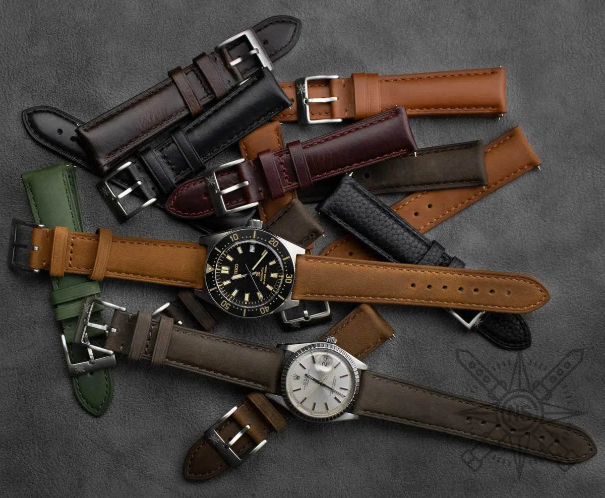 Two luxury wristwatches surrounded by a collection of colorful leather and textured watch straps on a grey surface.