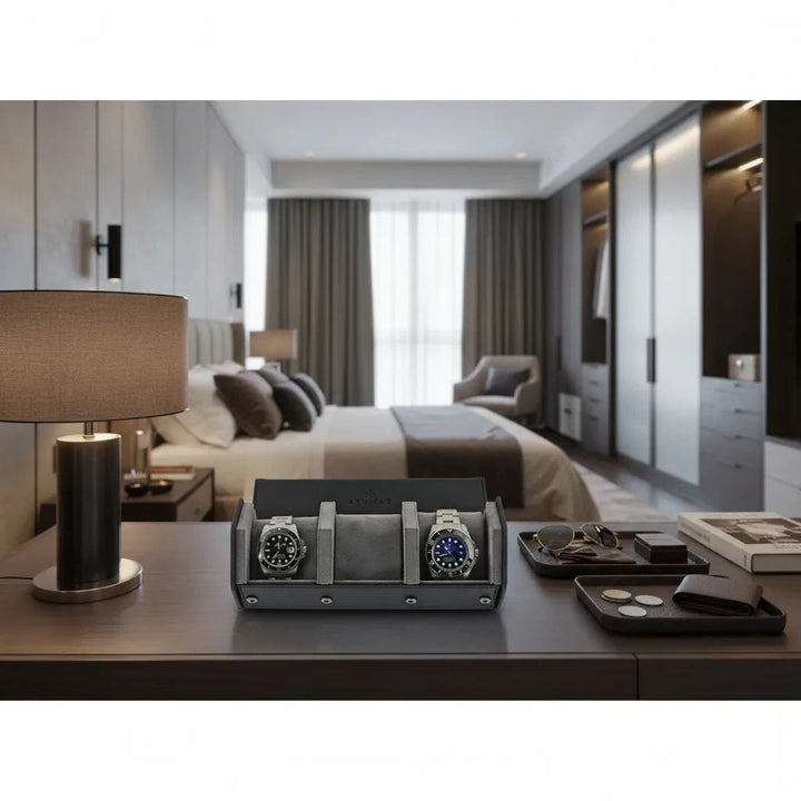 A modern bedroom with a lamp, eyeglasses, wallet, books, and the Aevitas Watch Winders 3 Watch Travel Case in black leather with grey lining (hexagon style) on a wooden desk; bed and large windows in the background.
