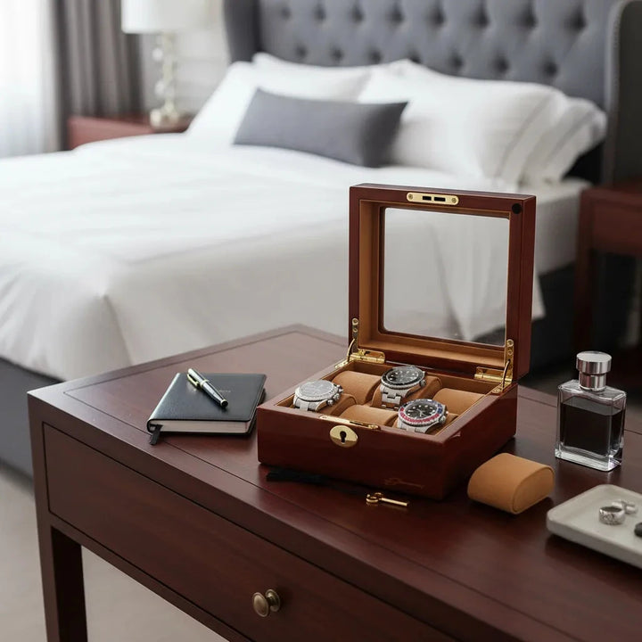The Aevitas 6 Watch Box in luxurious mahogany wood with camel lining sits on a bedside table with a notebook, pen, cologne bottle, and watch cushion. A neatly made bed with white linens and pillows completes the elegant scene.