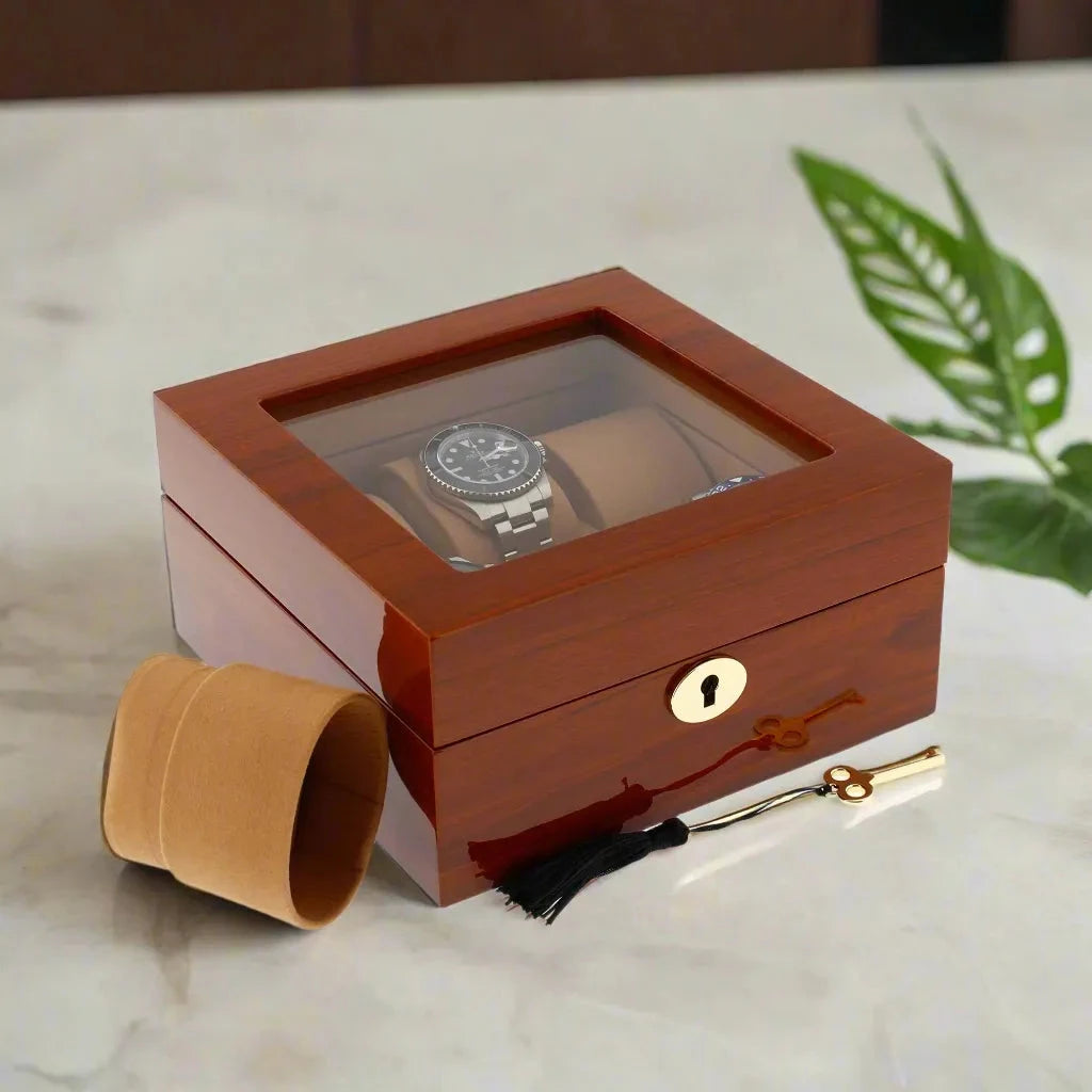 Showcasing the Aevitas 6 Watch Box in Luxurious Mahogany Wood with Camel Lining: a wristwatch displayed inside, a tan pillow and gold key with black tassel nearby. Green leaves and marble surface complete the elegant scene.