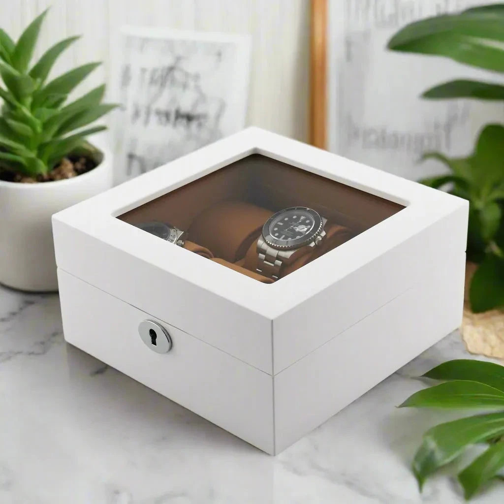 The Aevitas 6 Watch Box in Luxurious White Wood with Camel Lining displays wristwatches beneath its glass top, set on marble and surrounded by green potted plants and framed art.