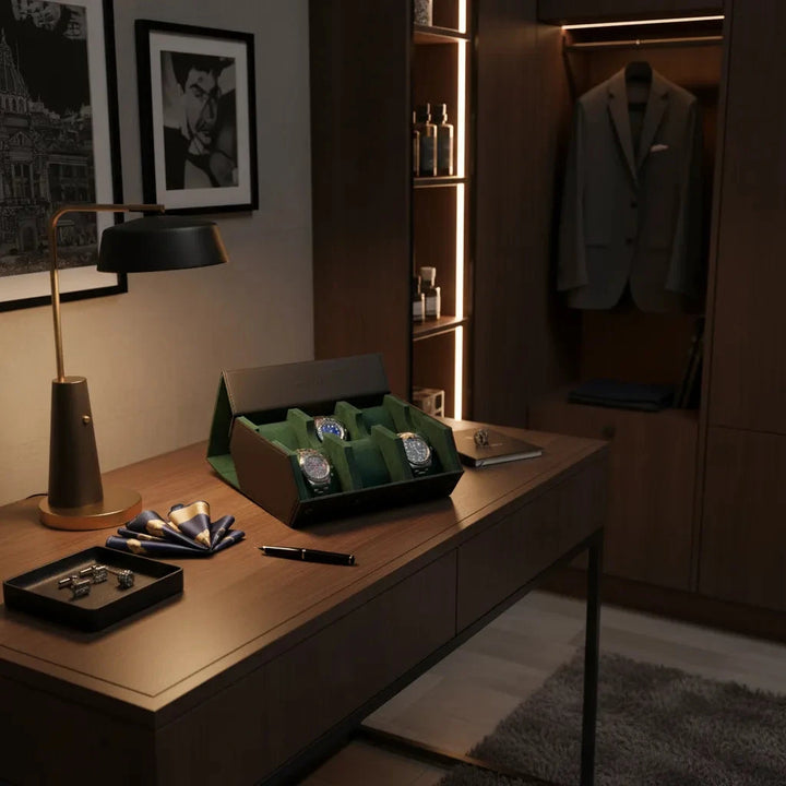 A stylish office features a wooden desk with a lamp, pens, cufflinks, pocket squares, and an open Aevitas Watch Winders 6 Watch Travel Case in Black Leather with Green Lining Hexagon Style displaying luxury watches. A gray suit hangs in a lit closet nearby.
