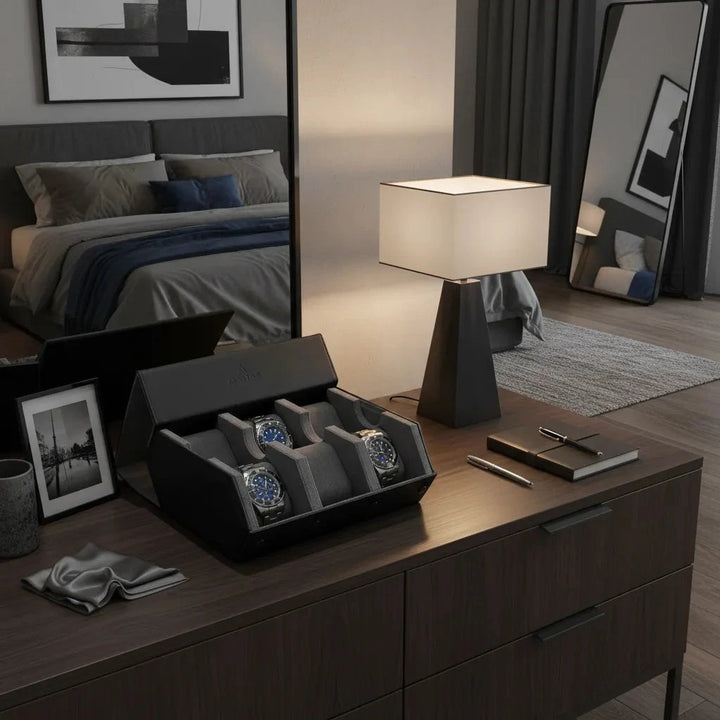 A modern bedroom features a bed, large mirror, nightstand, and on the wooden dresser—a lamp, photo frame, notepad, pen, cloth, and the Aevitas 6 Watch Travel Case in black leather with grey hexagon lining displaying four wristwatches.