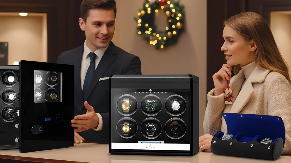 A man in a suit and a woman in a coat talk at a counter displaying luxury watch winders and a blue watch case, with holiday decorations in the background.