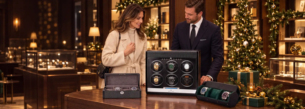 A man and a woman stand in a luxury watch winder boutique next to a large black 6-watch automatic watch conservory display case. The man wears a dark navy suit, and the woman wears a beige trench coat.