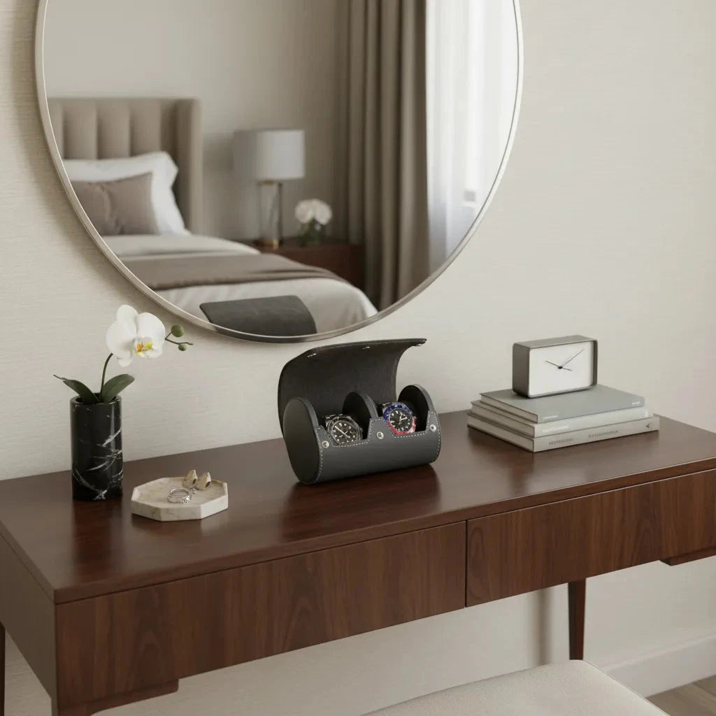 A wooden vanity with a round mirror displays the Aevitas Double Watch Roll in smooth grey leather with super soft lining holding two watches, a vase with a white orchid, jewelry on a tray, a clock, and stacked books; the bed is seen in the mirror.
