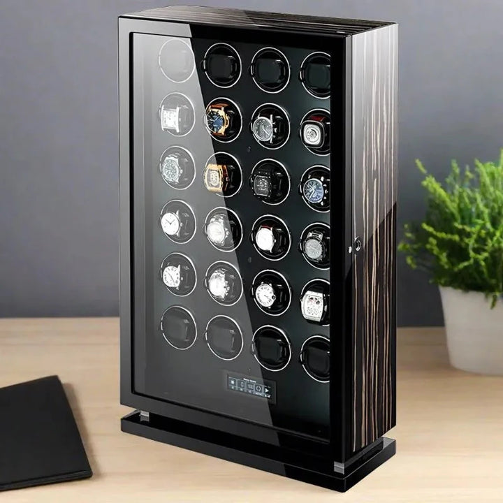 The Aevitas Premium 24 Watch Winder in striped ebony wood piano lacquer elegantly displays up to 24 watches in five rows and rests on a light wooden surface beside a green potted plant.