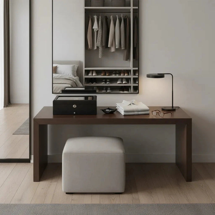 A modern, minimalist dressing area features a black oak table, beige ottoman, lamp, folded clothes, glasses, an Aevitas Premium Black Oak Veneer Watch Box for 10 Watches with Extra Storage Drawer, and jewelry box. A mirror reflects an open closet.