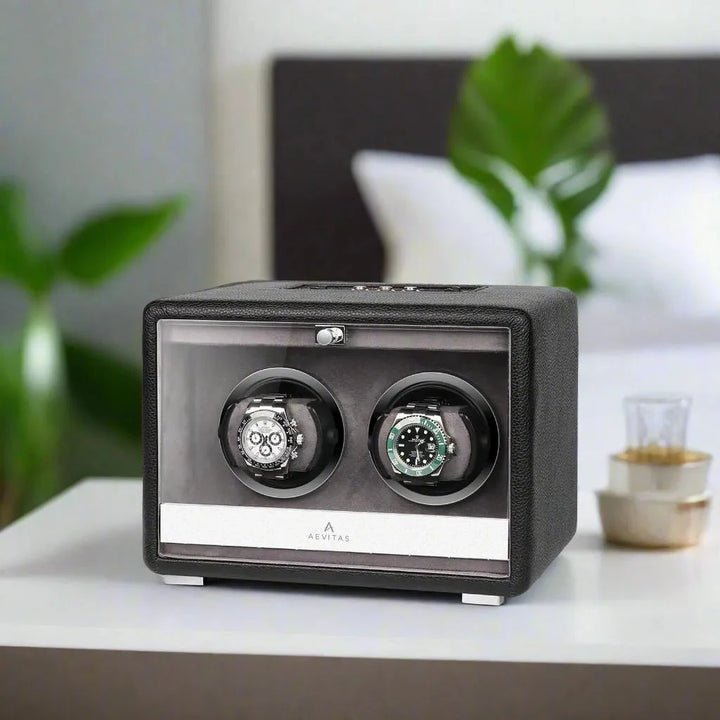 The Aevitas Premium Dual Watch Winder in black genuine leather sits atop a white bedside table, mains powered for quiet operation. Two luxury automatic watches rotate smoothly, with a bed and green plant softly blurred behind.