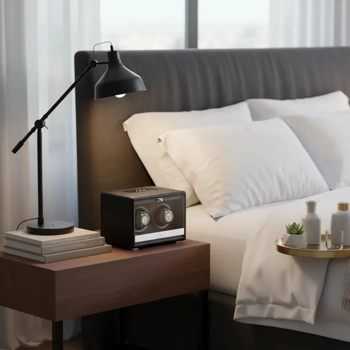 A modern bedroom with a neatly made bed, dark headboard, bedside table with lamp and books, and an Aevitas Premium Dual Watch Winder in black genuine leather. Sheer curtains let in natural light, enhancing the room’s stylish comfort.