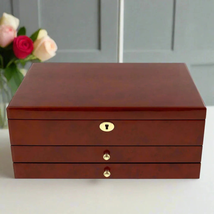 The Aevitas Watch Winders Premium Jewellery Box Extra Large Size, featuring a camphor finish and camel colour lining, sits on a white surface with a vase of pink and white roses before gray paneled doors in the background.