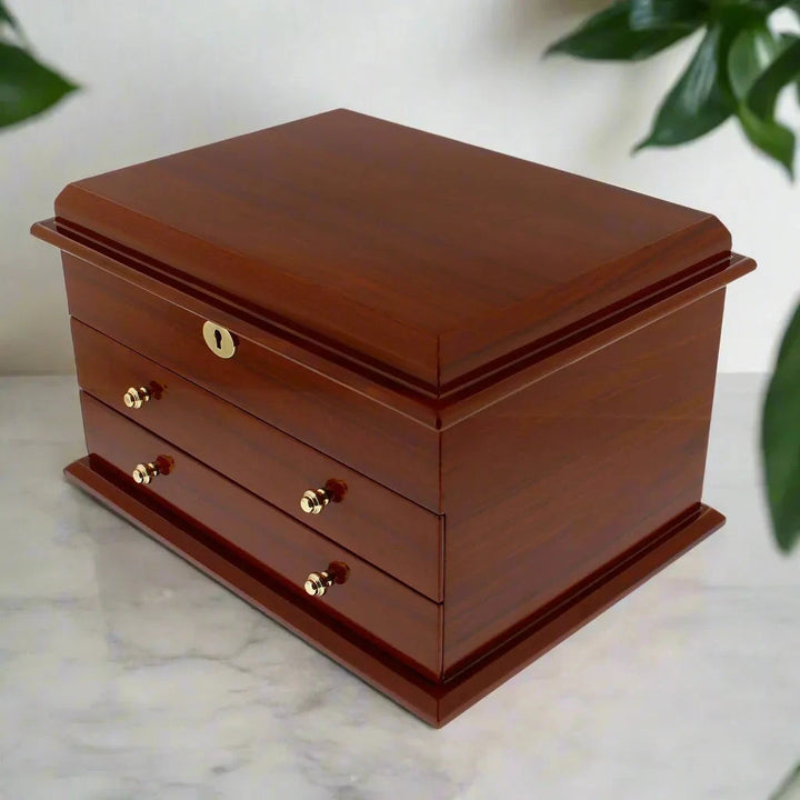 The Aevitas Premium Jewellery Box, Extra Large in mahogany wood with camel colour lining, features a hinged lid and two gold-knob drawers. Perfect for elegant jewellery storage, it sits beautifully on a white marble surface with green leaves behind.