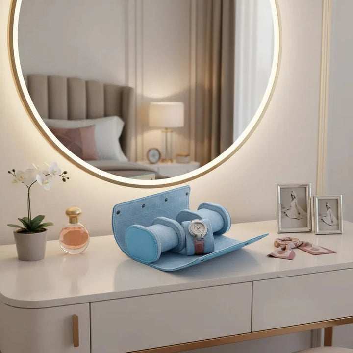 The Aevitas Premium Ladies 3 Watch Roll in Blue Saffiano Leather with Soft Lining holds a wristwatch on a white vanity, surrounded by perfume, an orchid, jewelry, and photos; the cozy bedroom reflected in the mirror adds elegance.