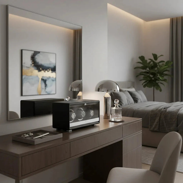 A modern bedroom features a wooden vanity with the Aevitas Premium Triple Watch Winder in black genuine leather, mains powered. A large mirror, decor accents, gray-bedded bed, potted plant, and abstract wall art complete this softly lit minimalist space.