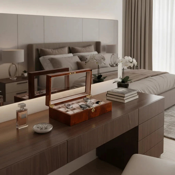 An Aevitas Watch Box for 12 Wrist Watches in Burl Walnut Wood sits atop a wooden dresser alongside perfumes, a jewelry dish, books, and potted orchids in a modern neutral bedroom with a neatly made bed and large mirror.