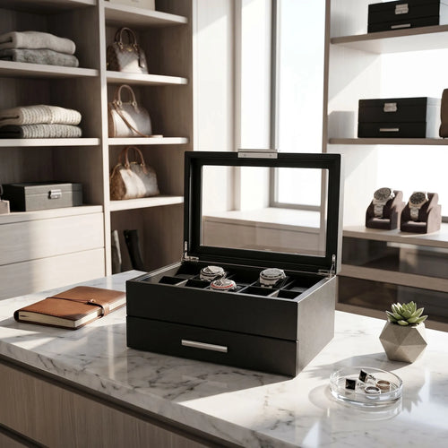 Black saffiano leather watch box with glass lid and drawer, displayed in a bright modern walk-in dressing room with elegant props.